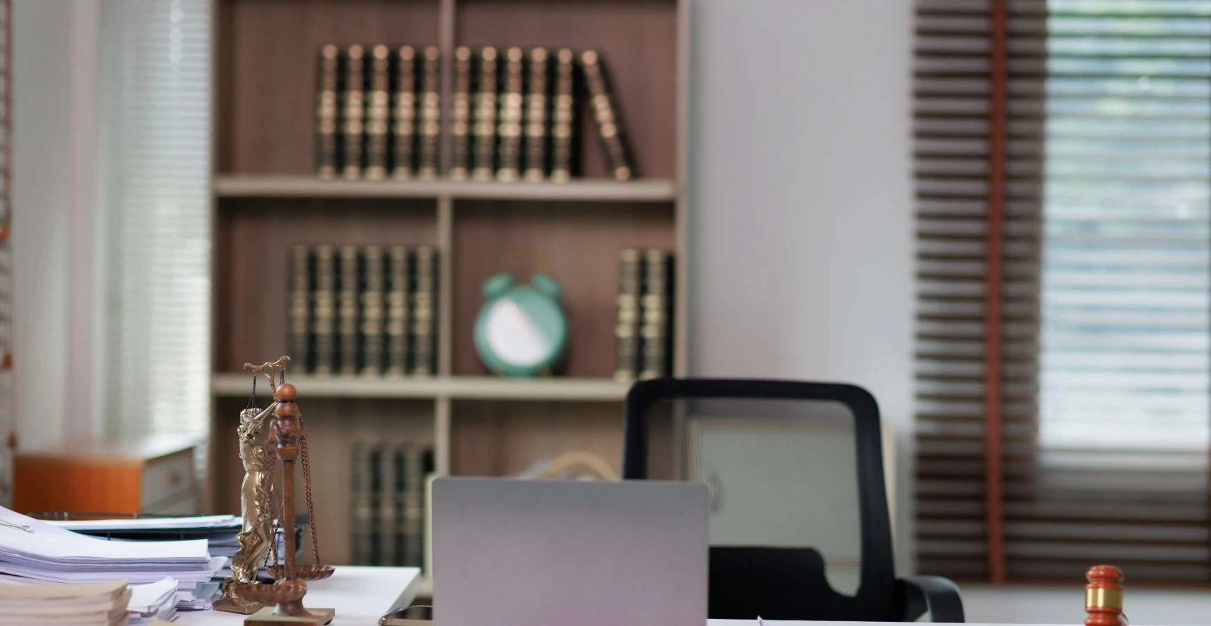 A vacant office desk featuring a laptop, stacks of paperwork, a scale of justice, and a background bookshelf, illustrating a legal firm providing compliance services.