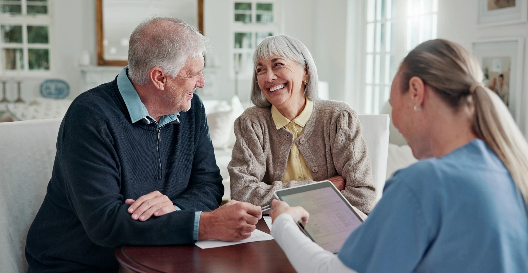 A happy senior man and woman smile during a consultation with a healthcare advisor or agent who is showing information on a digital tablet.