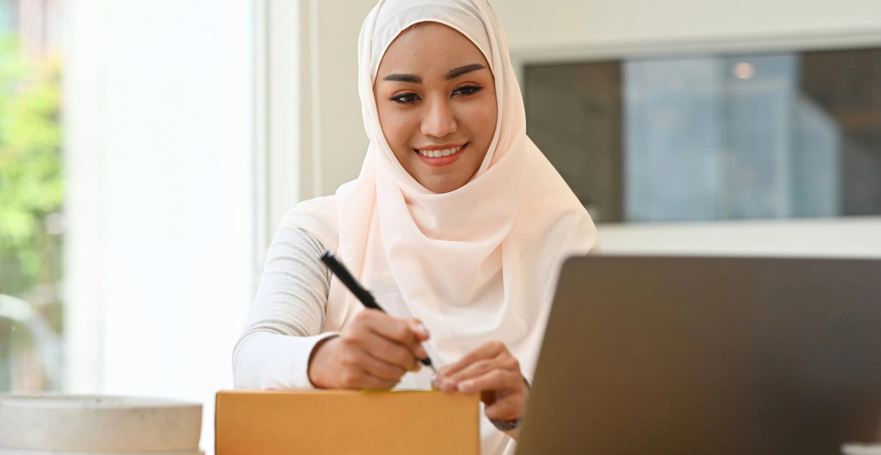 A smiling woman wearing a hijab processes orders on a laptop and writes on a cardboard box, showcasing the launching stage of a registered online shopping venture under an E-Commerce Business License.