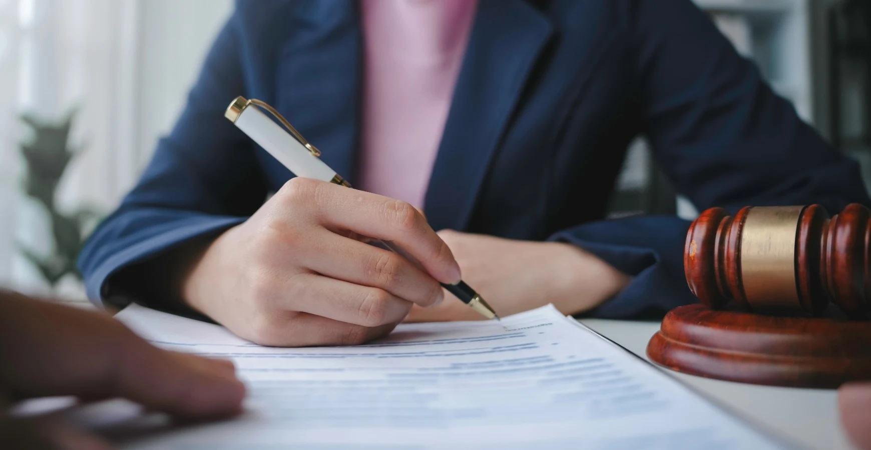 A legal professional in a dark blue blazer holds a white pen to sign an official contract next to a wooden gavel on a desk, illustrating formal Power of Attorney Services in the UAE.