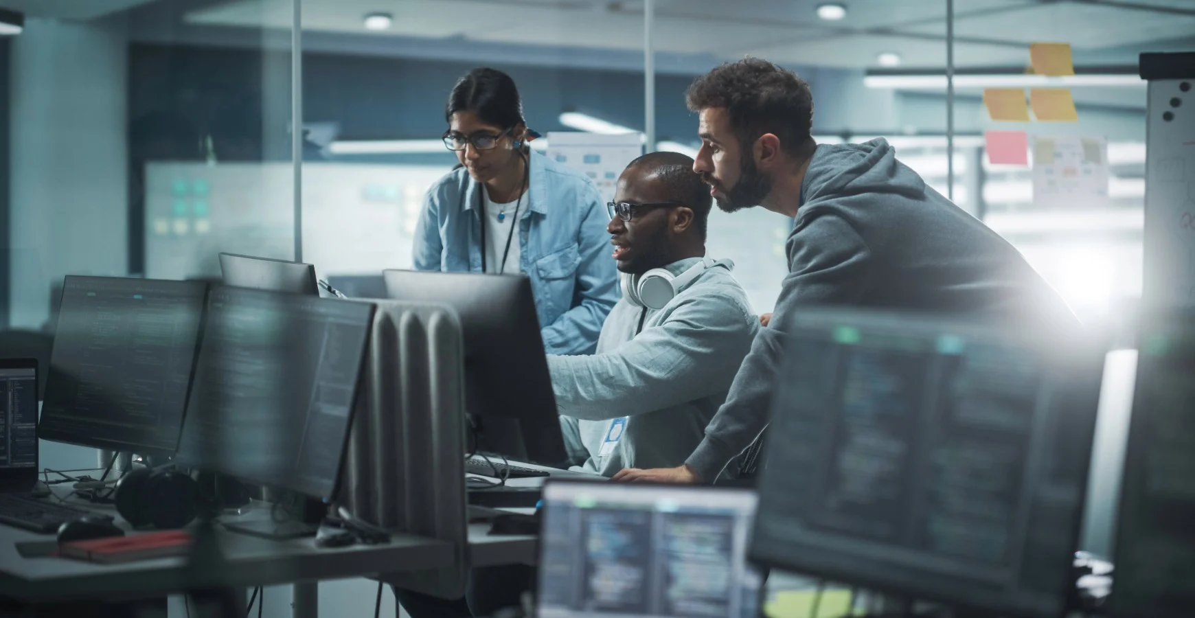 Three IT professionals—a woman and two men—work together in a modern office, analyzing code on multiple computer monitors.